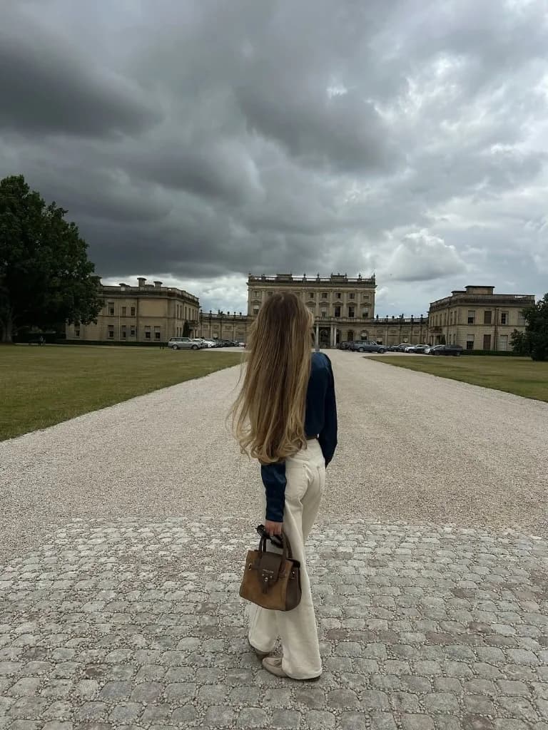 Junge Frau mit langen Haaren vor einem historischen Anwesen, weite Allee, bedeckter Himmel